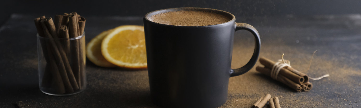 Eine schwarze Tasse auf dunklem Untergrund daneben liegen Zimtstangen