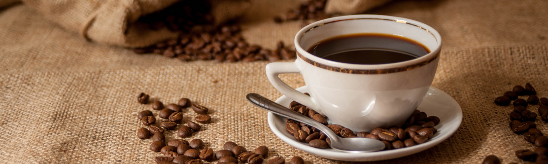 Eine Tasse mit Kaffee auf einem Unterteller mit einem Löffel und daneben liegen Kaffeebohnen