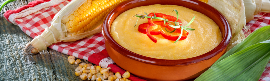 Schüssel mit Maisgrieß, garniert mit Paprika und Rosmarin, daneben frische Maiskolben und Maiskörner